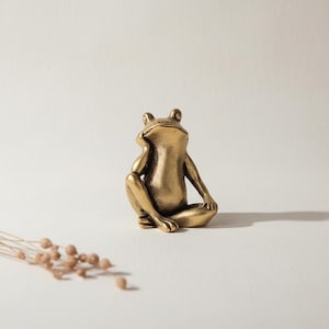 Puede incluir: Una figura de rana dorada en posición sentada, con la barbilla apoyada en la mano. La rana tiene detalles visibles y está sobre una superficie blanca. Unas cuantas ramas secas están en la esquina inferior izquierda.