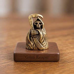 May include: A bronze-colored Grim Reaper figurine holding a scythe, mounted on a dark wooden base. The base is engraved with the words "Grim Reaper". The figurine has a detailed skull face and draped robe.