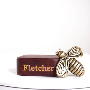 May include: A rectangular dark brown wooden block featuring the name Fletcher engraved in gold lettering on the front, accompanied by a metallic brass-toned bee ornament attached to the right side with detailed wing and body segment textures.