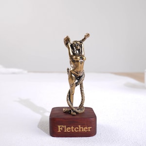 May include: A bronze-toned figurine of a person with arms raised, entwined by a snake, mounted on a rectangular dark wood base. The front of the wooden base features the name Fletcher engraved in a gold-colored serif font, centered on the face.