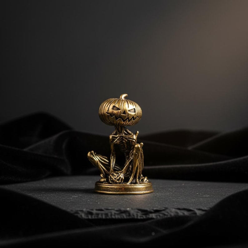May include: A small, metallic, brass-toned figurine depicting a seated, skeletal creature with a pumpkin head. The figure features a carved, grinning jack-o'-lantern face, thin limbs, and a textured, weathered finish, all mounted on a circular base.