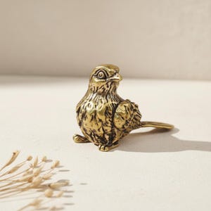 May include: A small, detailed brass bird figurine. The bird is in a seated position with its head turned slightly to the side. The brass has a warm, golden hue, and the bird's feathers are intricately textured.