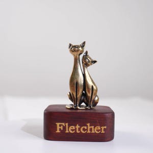 May include: A bronze-colored metal figurine of two cats sitting side-by-side, mounted on a rectangular dark brown wooden base. The word Fletcher is engraved in gold-colored lettering on the front face of the wooden block base.