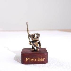 May include: A small bronze-coloured figurine of a stylised creature holding a tall staff, mounted on a rectangular dark wood base. The front of the wooden base features the name Fletcher engraved in a light-coloured, serif font.