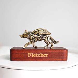May include: A metallic brass-toned wolf figurine with mechanical, segmented detailing on its back and legs, mounted on a rectangular dark reddish-brown wooden base with the name Fletcher engraved in a light gold font on the front face.
