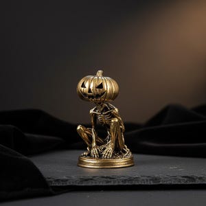 May include: A small, metallic gold-colored figurine depicting a crouching skeleton with a carved pumpkin head. The figure features detailed ribbing on the pumpkin and skeletal anatomy, set upon a circular base with a weathered, antique finish.