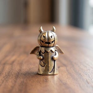 Peut inclure: Petite figurine de couleur laiton représentant un personnage sur le thème d'Halloween. La figurine a une tête en forme de citrouille avec un visage de lanterne sculpté, de petites cornes et des ailes. Sur une surface en bois, arrière-plan flou.