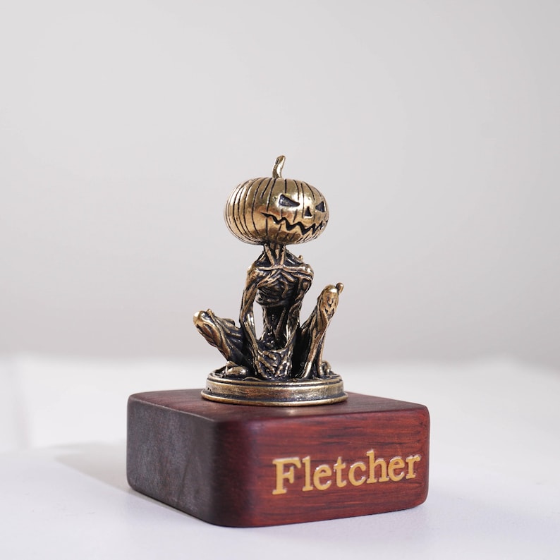 May include: A small bronze-colored metal figurine of a creature with a pumpkin head and a skeletal body, seated on a dark rectangular wooden base. The name Fletcher is engraved in gold lettering on the front face of the wooden base.