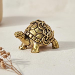 Op de afbeelding: Een kleine, gedetailleerde messing schildpadfiguur. De schildpad heeft een getextureerd schild, vier poten en een gedetailleerde kop. Een ketting van munten is over de rug van de schildpad gedrapeerd.