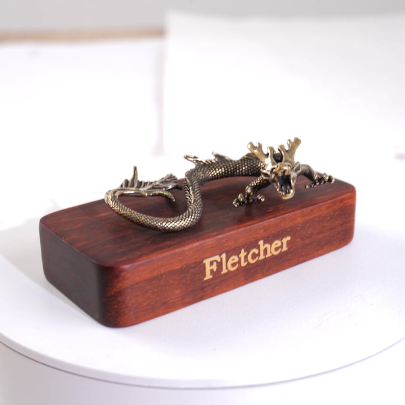 Op de afbeelding: Een rechthoekig donkerbruin houten blok met de naam Fletcher in goud gegraveerd op de voorzijde. Een metalen, bronskleurig drakenfiguurtje met gedetailleerde schubben en een open mond is bevestigd op de bovenzijde van het houten blok.
