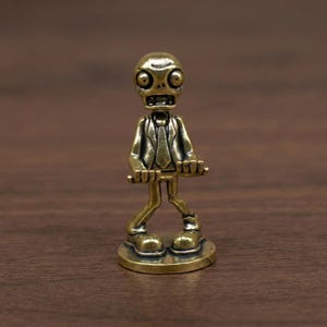 May include: A small, brass-coloured figurine of a zombie wearing a suit and tie. The zombie has large, round eyes and a wide, toothy grin. It stands on a small, circular base and holds its arms out in front of it.