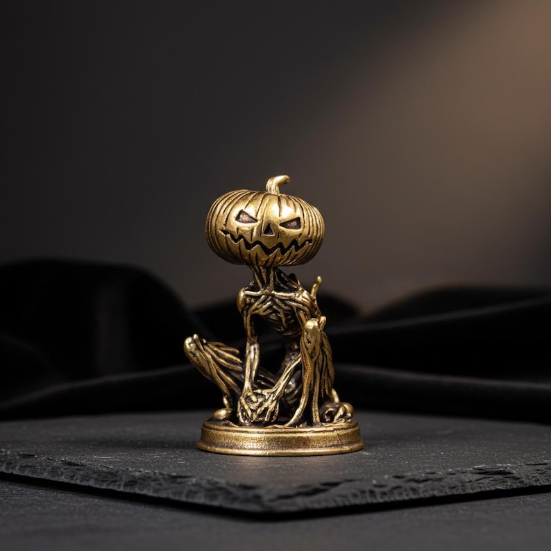 May include: A small, antique brass-colored figurine depicting a skeletal creature with a carved pumpkin head. The figure is in a seated position, featuring intricate, textured details on the body and limbs, all mounted on a circular base.