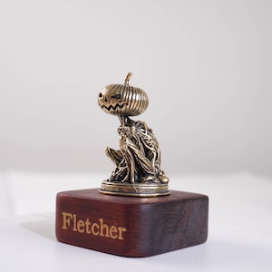 May include: A small bronze-toned figurine of a creature with a pumpkin head and a textured, vine-like body, mounted on a rectangular dark wood base. The word Fletcher is engraved in gold lettering on the front face of the wooden block.