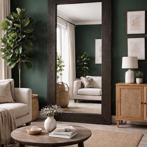 Puede incluir: Un gran espejo rectangular con un marco de madera oscura domina una escena de sala de estar. La habitaci&oacute;n presenta una paleta de colores neutros con una mesa de centro redonda, una alfombra beige y una pared verde. Los elementos decorativos incluyen una l&aacute;mpara blanca e impresiones bot&aacute;nicas enmarcadas.