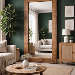 May include: A large, rectangular wooden-framed mirror dominates a room with a dark green wall. A round wooden coffee table, a white sofa, and a woven rug are in front of the mirror. Two framed botanical prints hang on the wall.