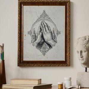 May include: A framed wall art print featuring a black and white ink illustration of two hands pressed together in prayer. The hands are centered within an ornate, decorative oval frame, with silhouettes of church steeples visible in the background.