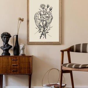 May include: A framed wall art print featuring a black and white ink illustration of two hands reaching towards each other, holding a thorny rose stem with one blooming rose and two rosebuds, set against a solid cream-coloured background.