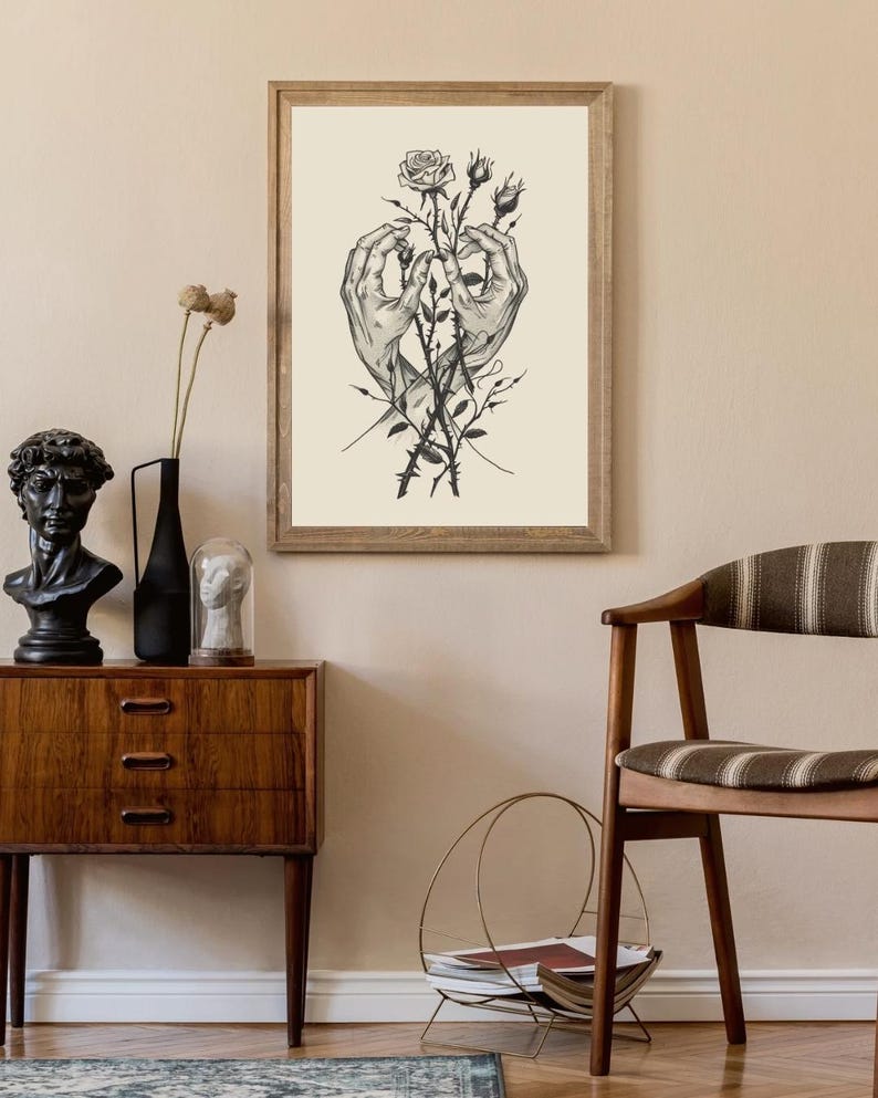 May include: A framed wall art print featuring a black and white ink illustration of two hands reaching towards each other, holding a thorny rose stem with one blooming rose and two rosebuds, set against a solid cream-coloured background.