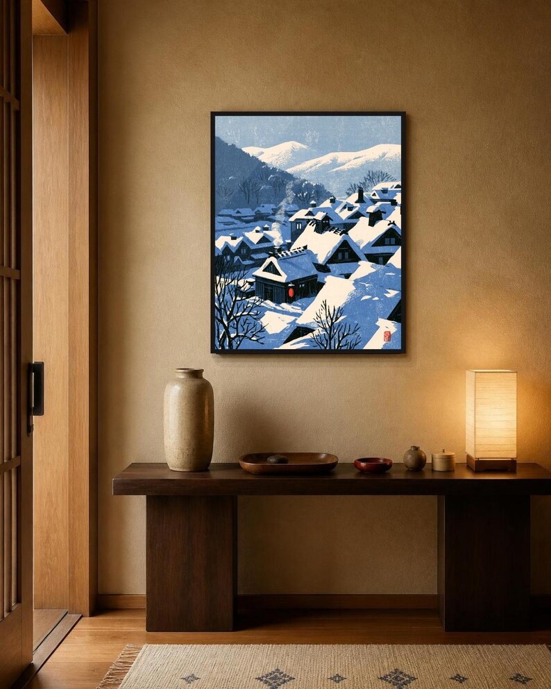 May include: A framed wall art print depicting a snowy village scene with dark-roofed houses covered in thick white snow, bare trees, and distant blue mountains under a clear sky. A single red lantern glows near a house with rising chimney smoke.