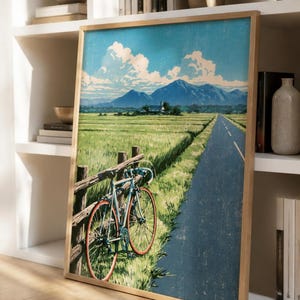 May include: A framed art print featuring a light blue bicycle leaning against a wooden fence beside a paved road. The landscape depicts a green field, a distant house, and blue mountains under a sky with white clouds, rendered in a textured style.