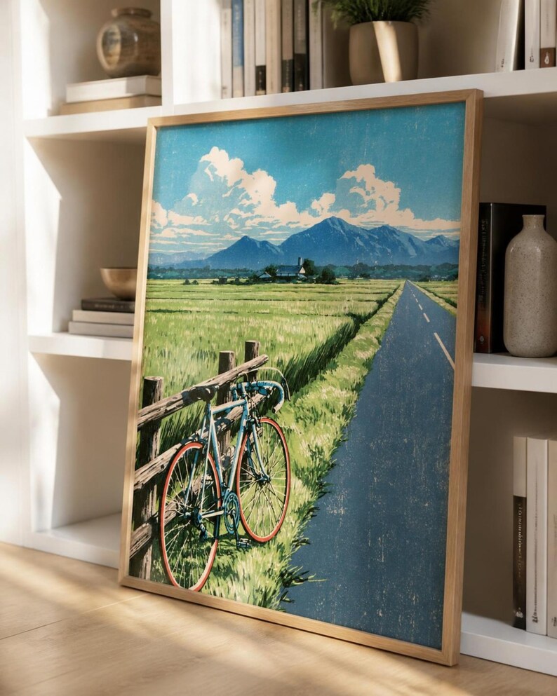 May include: A framed art print featuring a light blue bicycle leaning against a wooden fence beside a paved road. The landscape depicts a green field, a distant house, and blue mountains under a sky with white clouds, rendered in a textured style.