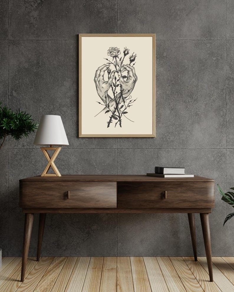 May include: A framed wall art print featuring a detailed black and white line drawing of two hands gently holding a rose stem with thorns. The illustration is set against a cream-coloured background and enclosed in a simple light wood frame.