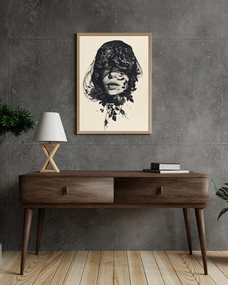 Op de afbeelding: Een ingelijste muurkunstprint met een zwart-witillustratie van een gezicht dat gedeeltelijk wordt bedekt door donkere rozen en vloeiende stof. Het kunstwerk staat op een lichte cr&egrave;mekleurige achtergrond in een eenvoudige houten lijst.