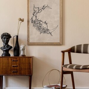 May include: A framed wall art print featuring a black line drawing of a crescent moon adorned with detailed roses and thorny vines on a light beige background. The artwork is displayed in a simple wooden frame, showcasing a botanical theme.