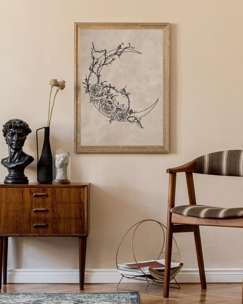 May include: A framed wall art print featuring a black line drawing of a crescent moon adorned with detailed roses and thorny vines on a light beige background. The artwork is displayed in a simple wooden frame, showcasing a botanical theme.