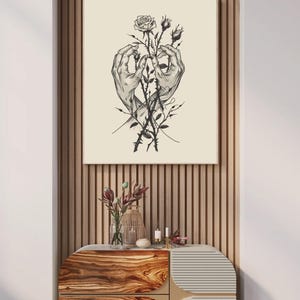 May include: A square wall art print featuring a black and white line drawing of two hands gently holding a rose stem with thorns and buds. The illustration is centred on a light cream background, presented in a thin frame on a slatted wall.