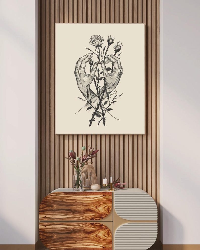 May include: A square wall art print featuring a black and white line drawing of two hands gently holding a rose stem with thorns and buds. The illustration is centred on a light cream background, presented in a thin frame on a slatted wall.