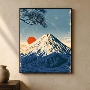 May include: A framed wall art print featuring a stylised illustration of a snow-capped mountain peak under a blue sky. A large, solid red sun sits behind the mountain, and a dark blue pine tree branch extends from the upper left corner.