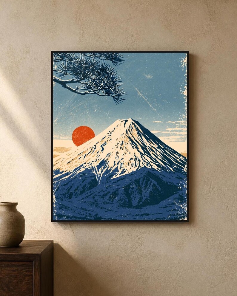 May include: A framed wall art print featuring a stylised illustration of a snow-capped mountain peak under a blue sky. A large, solid red sun sits behind the mountain, and a dark blue pine tree branch extends from the upper left corner.