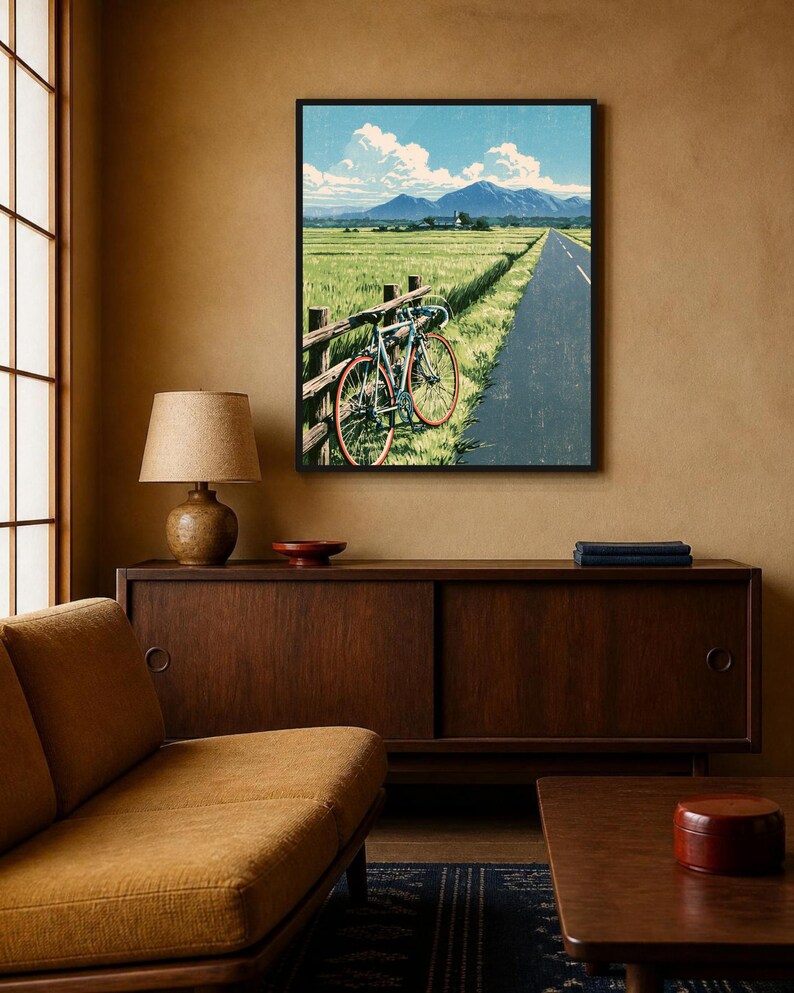 May include: A framed wall art print depicting a road bike leaning against a wooden fence beside a paved road. The scene features a green field, a distant mountain range under a blue sky with white clouds, and a small building in the background.