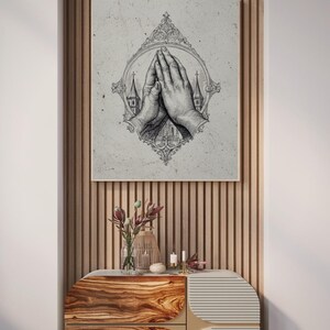 Op de afbeelding: Een vierkante muurkunstprint met een gedetailleerde zwart-witte inktillustratie van twee handen die in gebed zijn samengevouwen. De handen zijn gecentreerd in een sierlijk, ovaal frame met aan weerszijden kleine kerktorens.