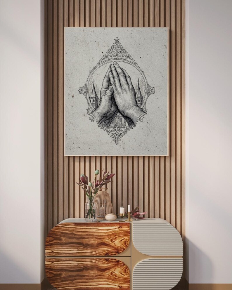 Op de afbeelding: Een vierkante muurkunstprint met een gedetailleerde zwart-witte inktillustratie van twee handen die in gebed zijn samengevouwen. De handen zijn gecentreerd in een sierlijk, ovaal frame met aan weerszijden kleine kerktorens.
