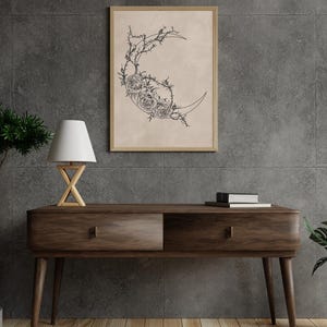 May include: A framed wall art print featuring a black line drawing of a crescent moon adorned with detailed roses and thorny vines on a light beige background. The artwork is displayed in a simple wooden frame hanging on a dark gray wall.
