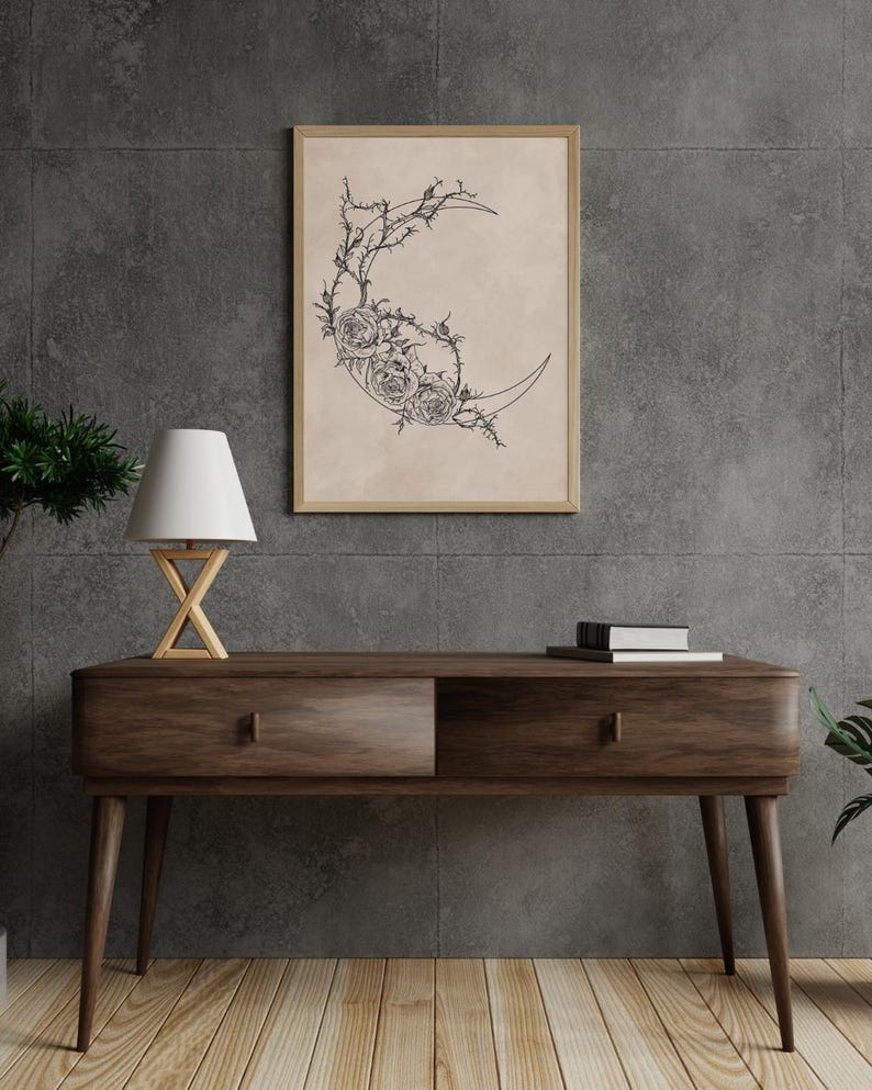 May include: A framed wall art print featuring a black line drawing of a crescent moon adorned with detailed roses and thorny vines on a light beige background. The artwork is displayed in a simple wooden frame hanging on a dark gray wall.