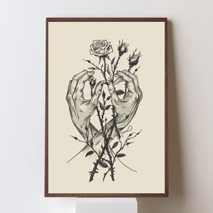 May include: A framed wall art print featuring a black and white ink illustration of two hands gently cradling a cluster of rose stems with thorns and buds. The artwork is set against a solid cream-coloured background within a dark brown frame.