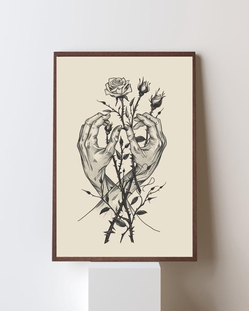 May include: A framed wall art print featuring a black and white ink illustration of two hands gently cradling a cluster of rose stems with thorns and buds. The artwork is set against a solid cream-coloured background within a dark brown frame.