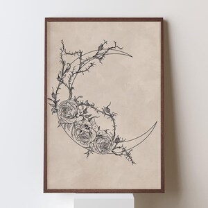 May include: A framed wall art print featuring a black line drawing of a crescent moon intertwined with thorny rose vines and three blooming roses on a solid beige background. The artwork is enclosed in a thin, dark brown wooden frame.