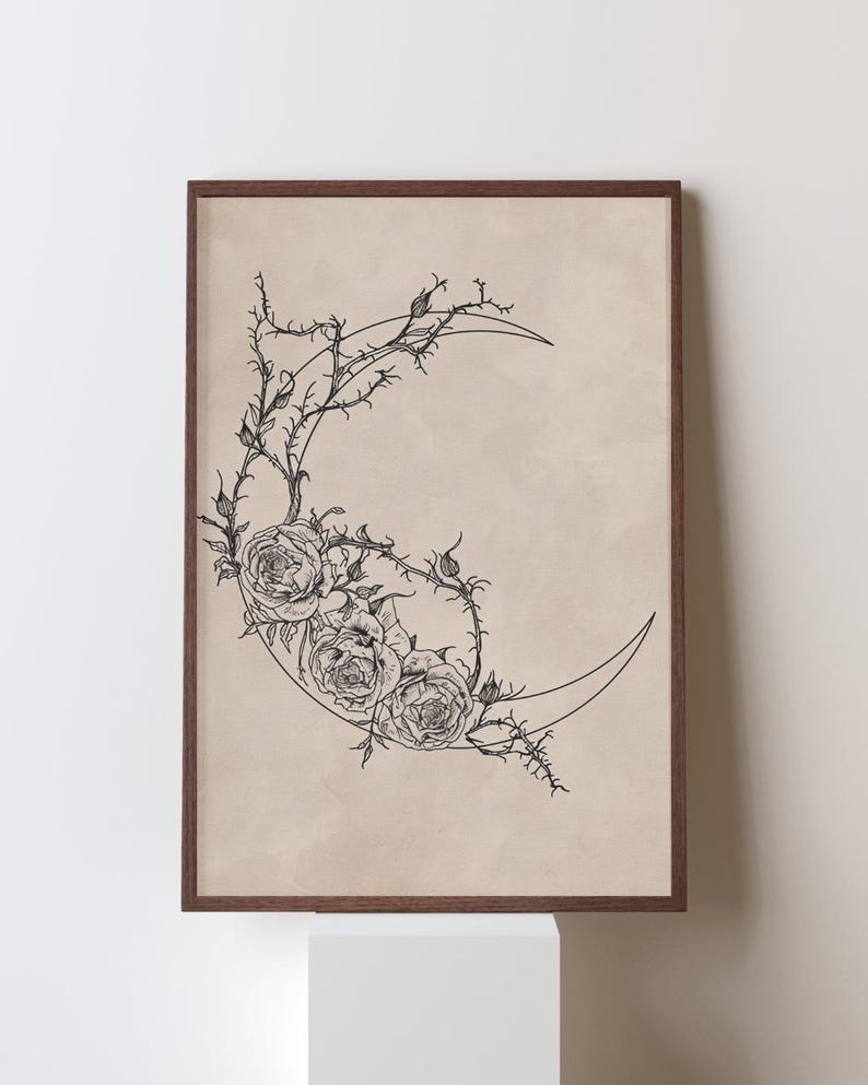 May include: A framed wall art print featuring a black line drawing of a crescent moon intertwined with thorny rose vines and three blooming roses on a solid beige background. The artwork is enclosed in a thin, dark brown wooden frame.