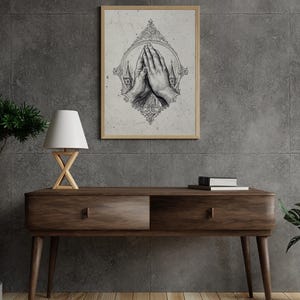 Op de afbeelding: Een ingelijste wandkunstprint met een gedetailleerde zwart-witillustratie van twee handen die in gebed zijn samengevouwen, omsloten door een sierlijk decoratief frame met kleine kerktorens aan weerszijden, tegen een lichte, getextureerde achtergrond.