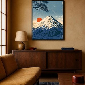 May include: A framed wall art print featuring a stylised illustration of a snow-capped mountain peak against a light blue sky. A vibrant red sun sits to the left of the mountain, with dark pine tree branches visible in the upper left corner.