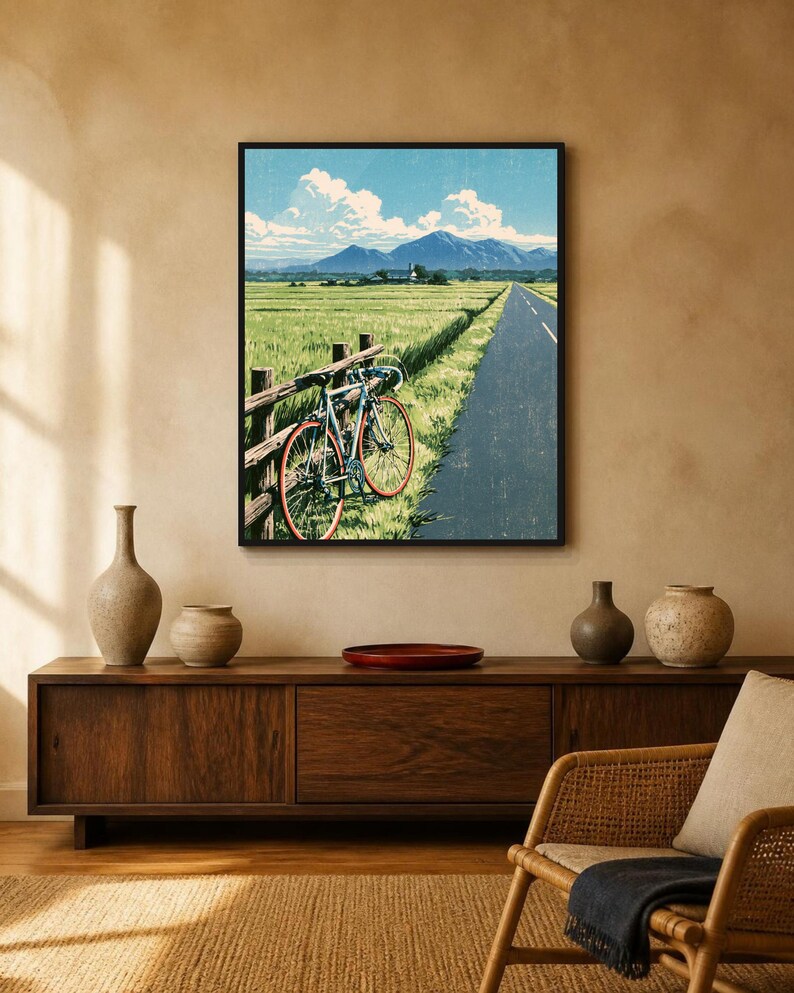 May include: A framed wall art print depicting a road bike leaning against a wooden fence beside a paved road. The scene features a green field, a distant mountain range under a blue sky with white clouds, and a textured, illustrated aesthetic.