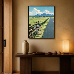 May include: A framed wall art print depicting a landscape scene with a bicycle leaning against a wooden fence beside a paved road. The background features a green field, distant blue mountains, and a bright blue sky with white clouds.
