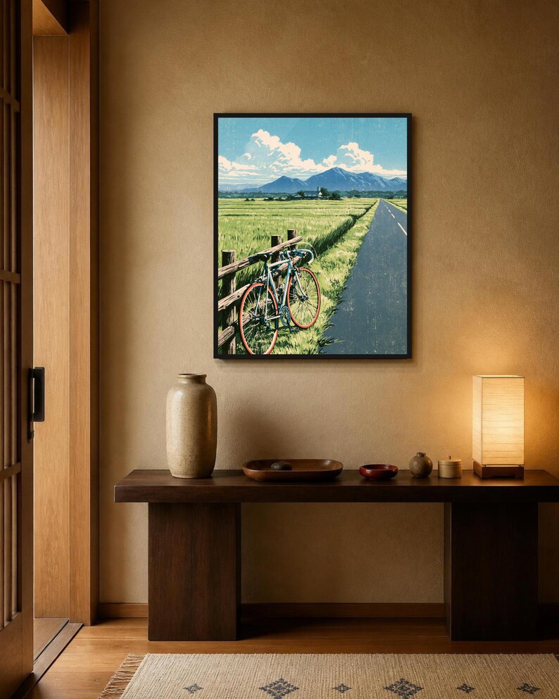 May include: A framed wall art print depicting a landscape scene with a bicycle leaning against a wooden fence beside a paved road. The background features a green field, distant blue mountains, and a bright blue sky with white clouds.