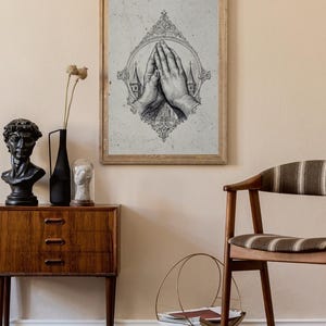 Op de afbeelding: Een ingelijste wandkunstprint met een gedetailleerde zwart-witillustratie van twee handen die in gebed zijn samengevouwen, omsloten door een sierlijke, decoratieve ovale lijst met kerktorens zichtbaar op de achtergrond.