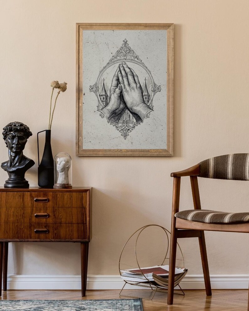 Op de afbeelding: Een ingelijste wandkunstprint met een gedetailleerde zwart-witillustratie van twee handen die in gebed zijn samengevouwen, omsloten door een sierlijke, decoratieve ovale lijst met kerktorens zichtbaar op de achtergrond.