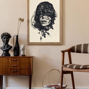 Op de afbeelding: Een ingelijste kunstdruk met een zwart-witillustratie van een gezicht dat gedeeltelijk wordt bedekt door een sluier van donkere rozen en bladeren. Het kunstwerk zit in een lichtgekleurde houten lijst en toont gedetailleerde florale vormen.